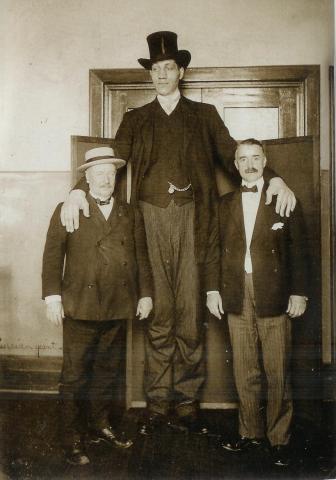 The 'Russian Giant' stands with his hands on the shoulders of two men in suits, whose heads reach only to the Russian man's waist. The men in suits each have moustaches. The Russian man wears a top hat and tuxedo with tails. The chain of a pocket watch is visible where his suit jacket is open. There is a set of double doors behind the men, and the Russian man is clearly taller than this doorway.